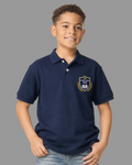 A young boy wearing a navy blue polo shirt with a school crest on the left chest, paired with blue jeans, standing against a light gray background.